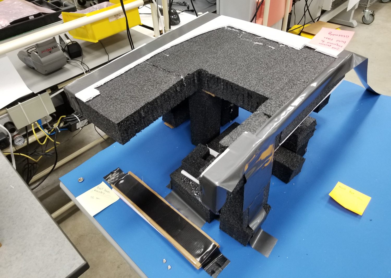 A full size mock-up of a machine enclosure cut from gray foam and sheet metal, standing on a workbench beside the real components.