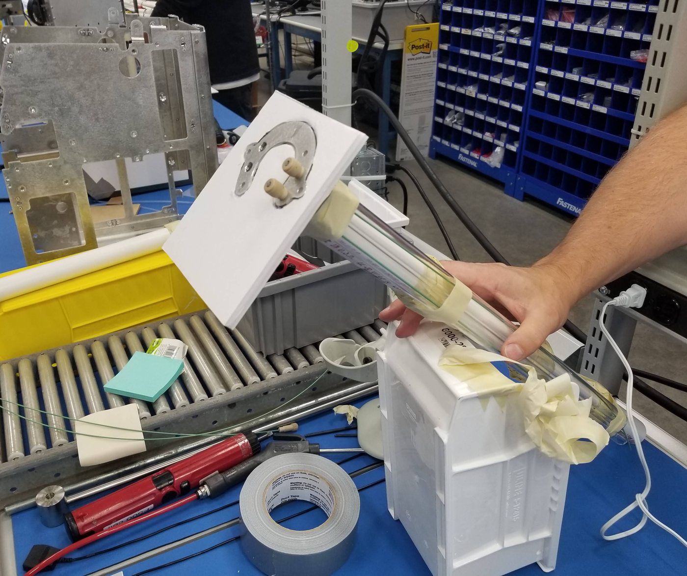 A hand holding a foam and cardboard mock-up of a sub-assembly above a workbench lined with tools and parts bins.
