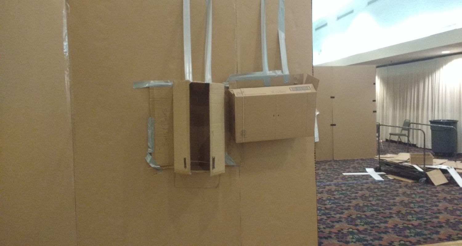 A full-scale cardboard mock-up of a room interior, with a taped fixture on the wall and further cardboard walls beyond.