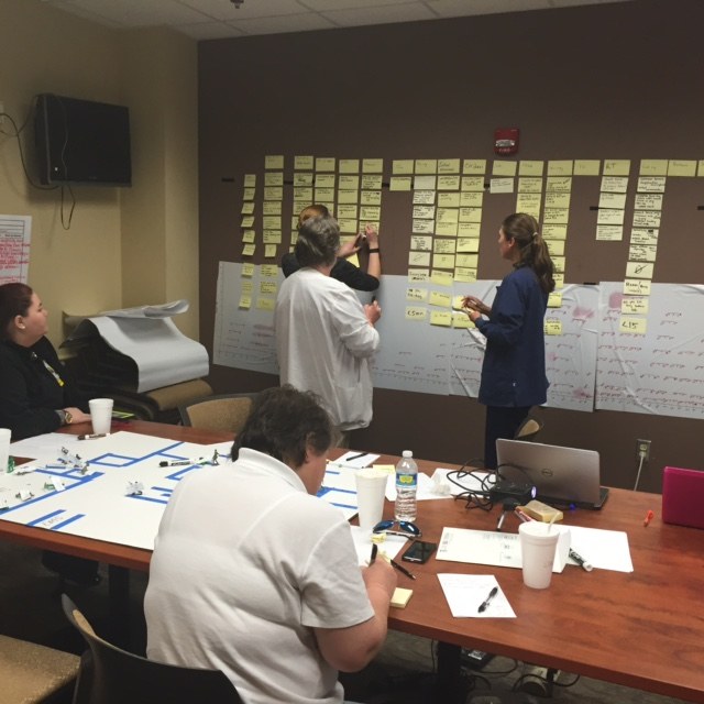 Two people standing at a wall covered in rows of sticky notes under printed column headings, adding another note, while colleagues work at a table in front of them.
