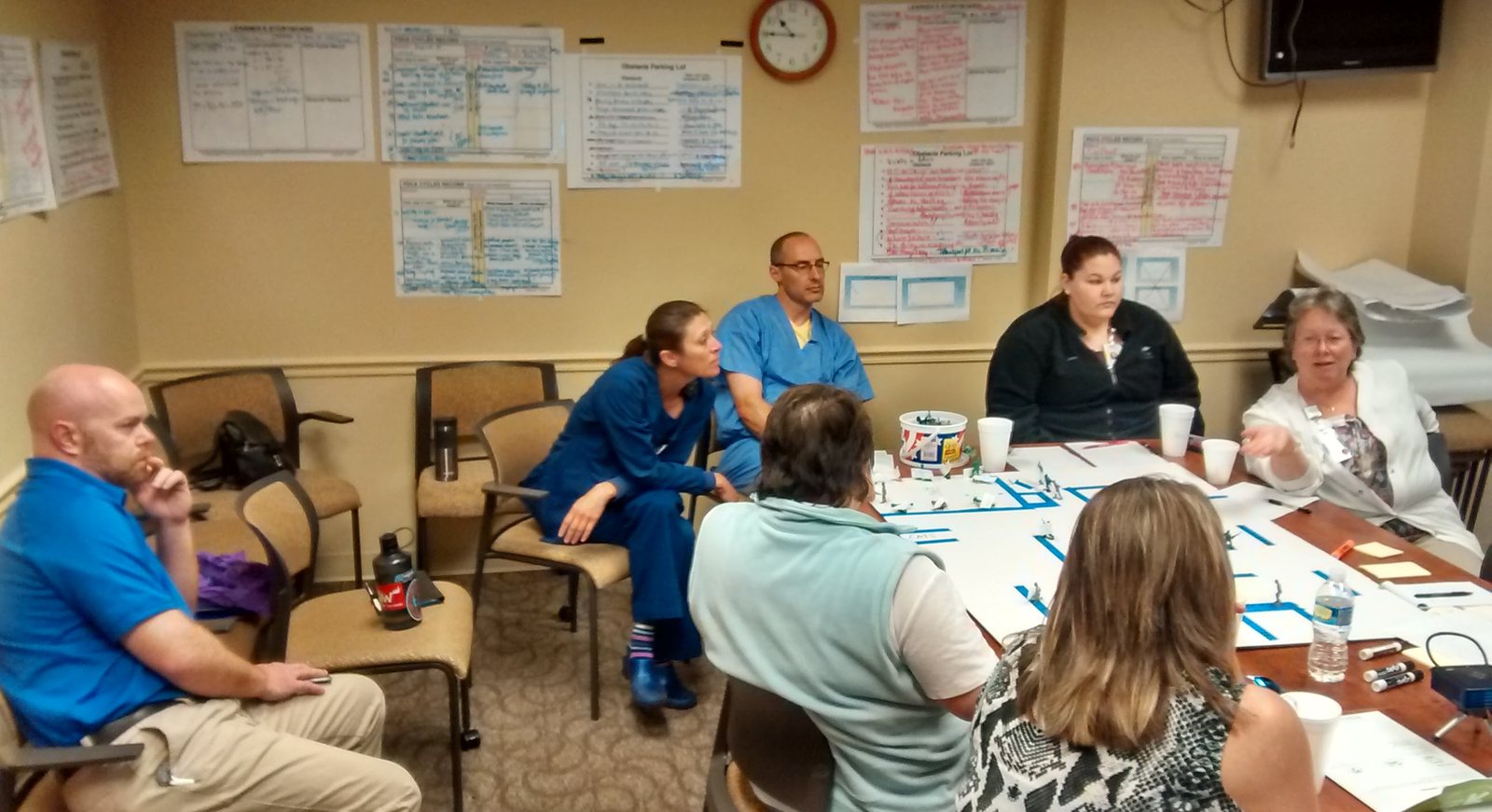 Clinical staff around a conference table, some in scrubs, working over a proposed department layout taped out in blue on butcher paper with small figures standing in the rooms. Learner's storyboards, obstacle parking lists and PDCA cycles records are pinned to the wall behind them.