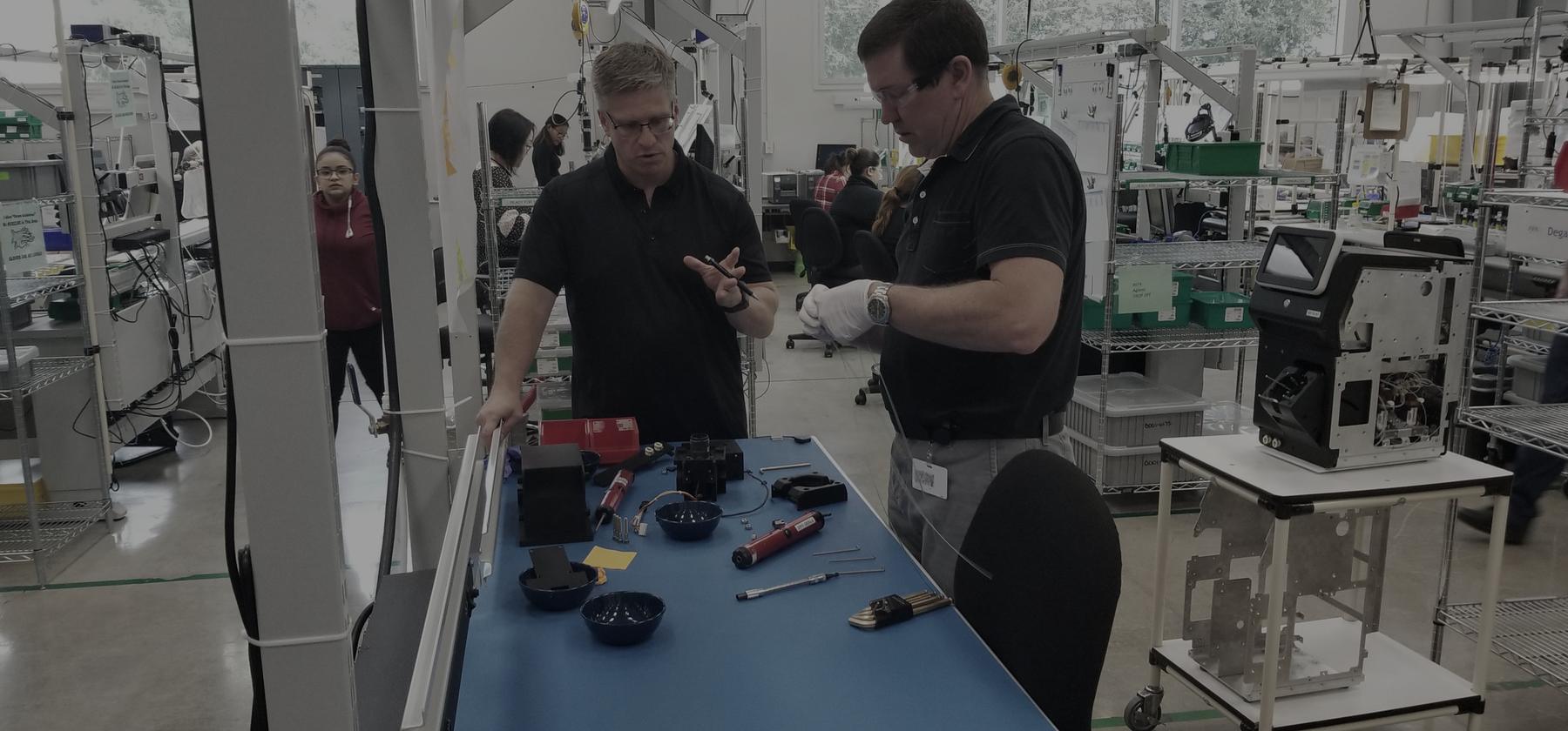 Two people standing at a workbench on a production floor, heads together over a part, with assembly stations and parts bins running away behind them.